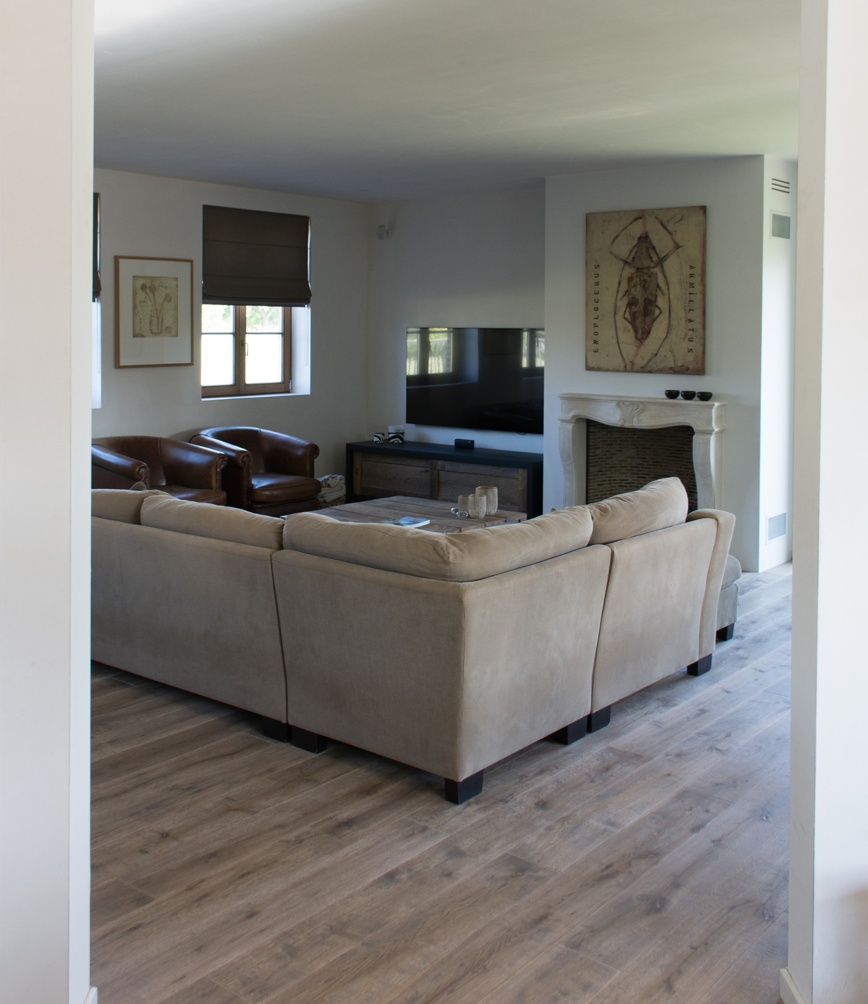 Salon z drewnianą podłogą ułożoną w jodełkę, beżowa narożna sofa, skórzane fotele, kominek z marmurową obudową i obrazem owada na ścianie.