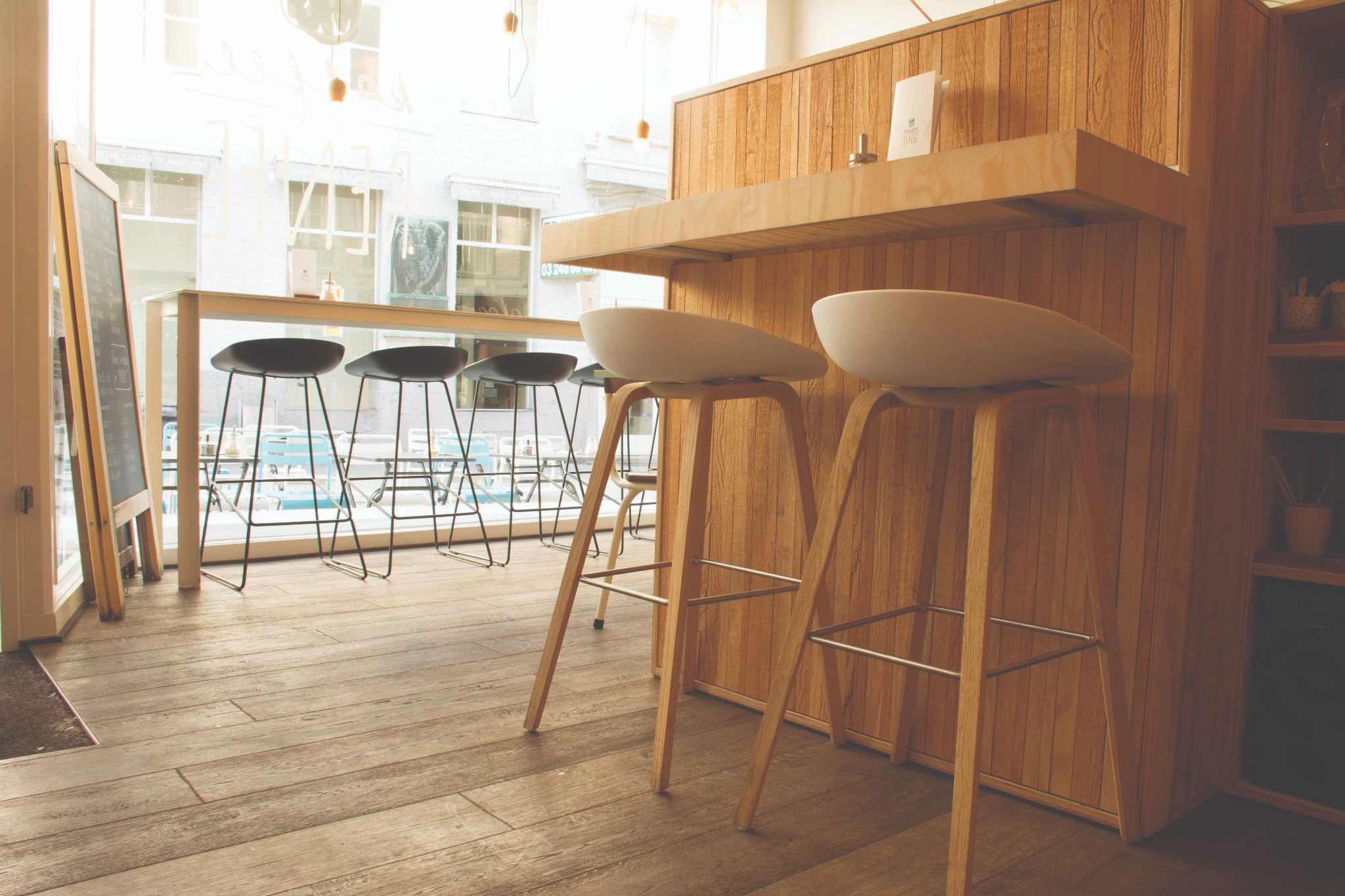 Wnętrze kawiarni z drewnianą podłogą, barowymi stołkami i ścianą wykończoną panelami z jasnego drewna.