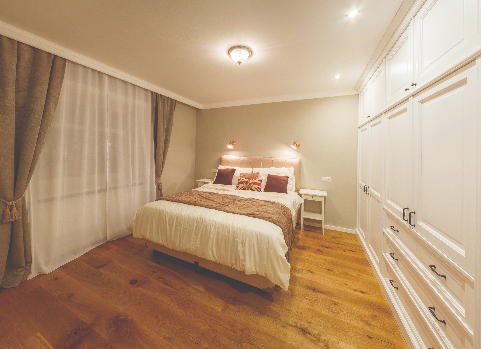 Jasna sypialnia z parkietem w odcieniach brązu, beżowymi zasłonami i białymi meblami w zabudowie.