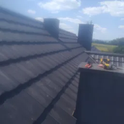 Ciemnoszary dach z dachówki ceramicznej z dwoma kominami, w tle zielony krajobraz i błękitne niebo z chmurami, na kominie leżą narzędzia: kombinerki i wkrętarka.