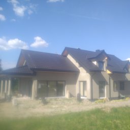 PRB DACHOVIA S.C - Nowo wybudowany, parterowy dom z ciemną dachówką, jeszcze w trakcie wykończenia elewacji, z widocznym pracownikiem w pomarańczowych spodniach po prawej stronie.