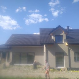 PRB DACHOVIA S.C - Nowoczesny dom jednorodzinny z ciemną, metalową dachówką, widoczny pracownik w pomarańczowych spodniach na tle częściowo wykończonej elewacji i błękitnego nieba z chmurami.