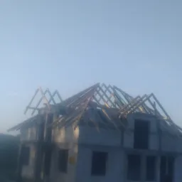 Surowy stan domu w budowie z widoczną drewnianą konstrukcją dachu, w tle zieleń drzew w porannym, zamglonym świetle.