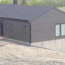 Nowoczesny dom szkieletowy z ciemną elewacją i dachem z dach&oacute;wki, widok z zewnątrz na nier&oacute;wny teren.