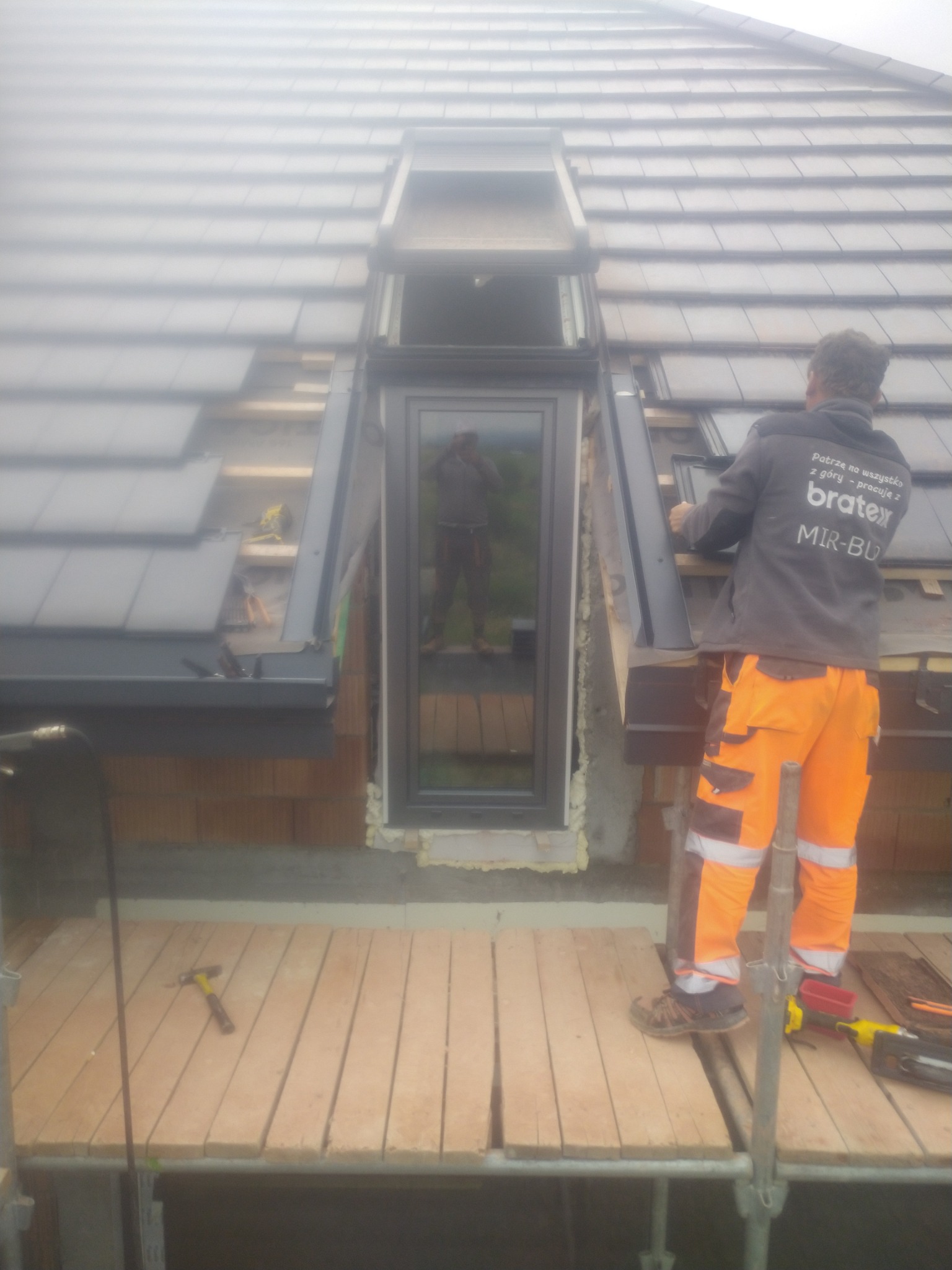 Montaż okna dachowego na pokrytym szarą dachówką dachu, widoczny dekarz w pomarańczowych spodniach i kurtce z logo Bratex, narzędzia na rusztowaniu.