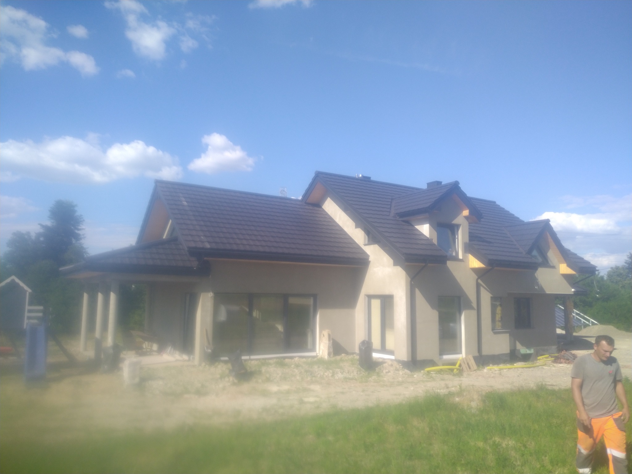Nowo wybudowany, parterowy dom z ciemną dachówką, jeszcze w trakcie wykończenia elewacji, z widocznym pracownikiem w pomarańczowych spodniach po prawej stronie.