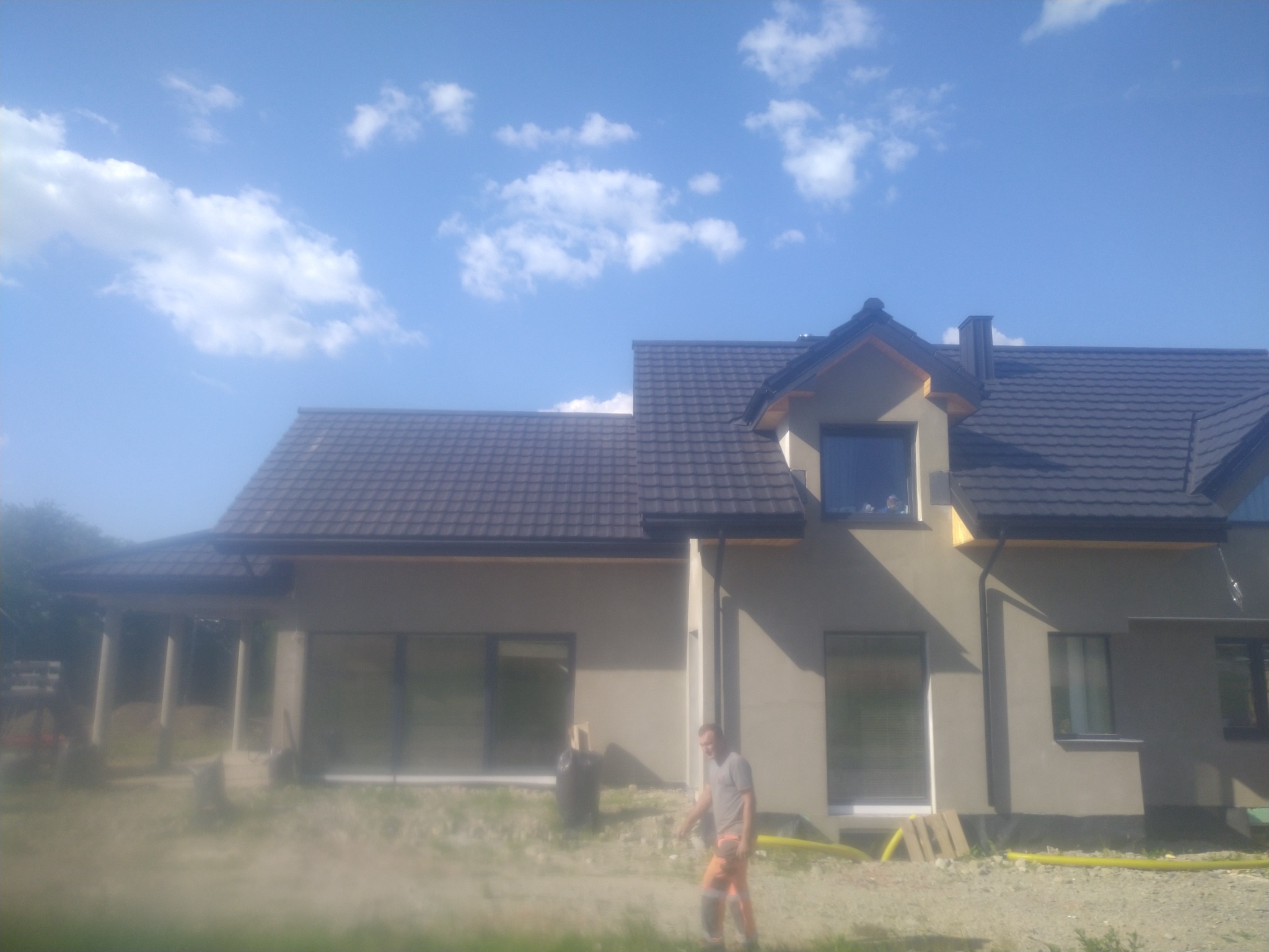 Nowoczesny dom jednorodzinny z ciemną, metalową dachówką, widoczny pracownik w pomarańczowych spodniach na tle częściowo wykończonej elewacji i błękitnego nieba z chmurami.