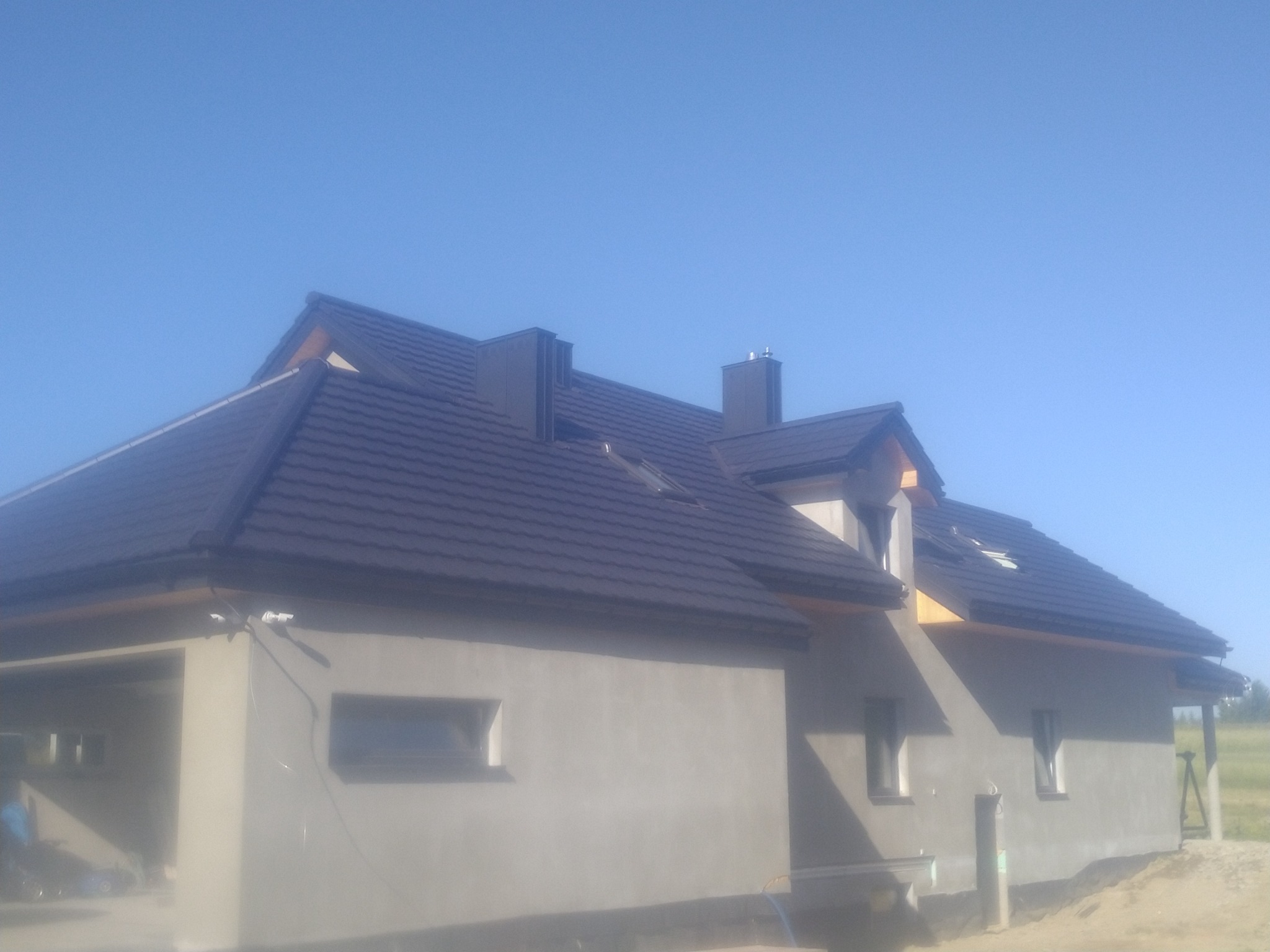 Dom jednorodzinny w trakcie budowy z ciemnym dachem z dachówki ceramicznej i widocznymi obróbkami blacharskimi, oświetlony jasnym słońcem, z nieotynkowaną elewacją i elementami wykończeniowymi...