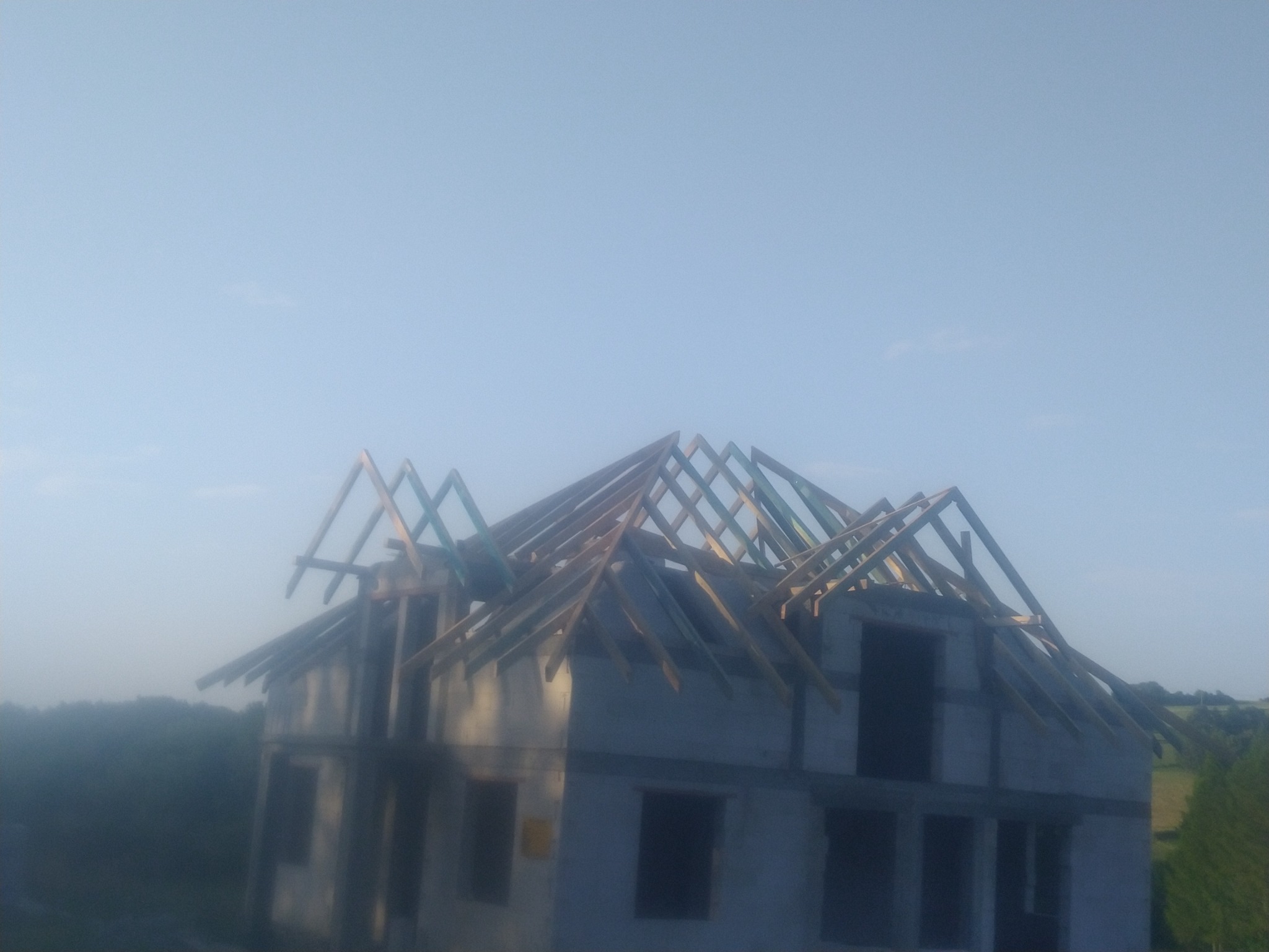 Surowy stan domu w budowie z widoczną drewnianą konstrukcją dachu, w tle zieleń drzew w porannym, zamglonym świetle.