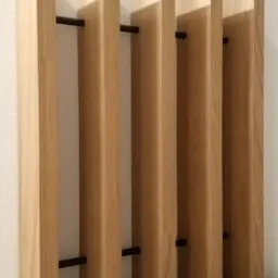 Drewniane lamele ścienne w jasnym odcieniu, przymocowane do ściany za pomocą czarnych prętów, ułożone na szarej, teksturowanej powierzchni.