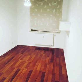 Pusty pokój z świeżo położoną, błyszczącą, ciemnobrązową podłogą z paneli, białe ściany, okno z zasłoniętą roletą, widoczne światło górne.