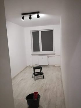 Pomalowane na biało wnętrze pokoju z jasną podłogą, czarnym stołkiem i wiadrem z czerwonym akcentem w rogu, oświetlone lampą sufitową z trzema reflektorami.