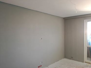 Świeżo pomalowane na szaro wnętrze pokoju z widocznymi gniazdkami elektrycznymi i fragmentem okna.