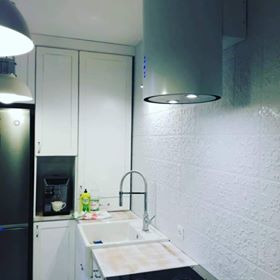 Jasna kuchnia z białymi szafkami, okapem cylindrycznym i strukturalną ścianą z płytek. Widoczny ekspres do kawy i zlewozmywak z chromowaną baterią.