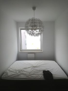 Pusta sypialnia z białymi ścianami, łóżkiem z pogniecioną białą pościelą i nowoczesną lampą w kształcie kuli wiszącą pod sufitem, widok z góry na okno z widocznym budynkiem na zewnątrz.