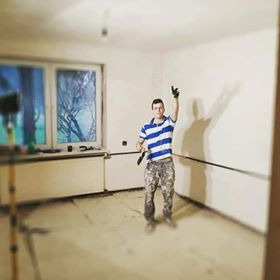 Młody mężczyzna w ubraniach roboczych z pędzlem w ręku, stojący w pustym, częściowo odmalowanym pokoju z widocznym grzejnikiem i oknem z widokiem na drzewa, wskazujący palcem na świeżo pomalowany...