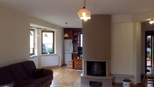 Jasne wnętrze salonu z kominkiem w odcieniach beżu i brązu, widok na okna i aneks kuchenny z białą lodówką, drewniana podłoga i sofa z poduszką.