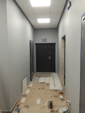 Korytarz w szarych odcieniach po świeżym malowaniu, widoczne zabezpieczone podłogi i przygotowane wejście do pomieszczenia bez drzwi.