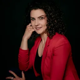 Uśmiechnięta kobieta z kręconymi włosami w czerwonej marynarce, opierająca brodę na dłoni, z pierścionkiem na palcu, na ciemnym tle.