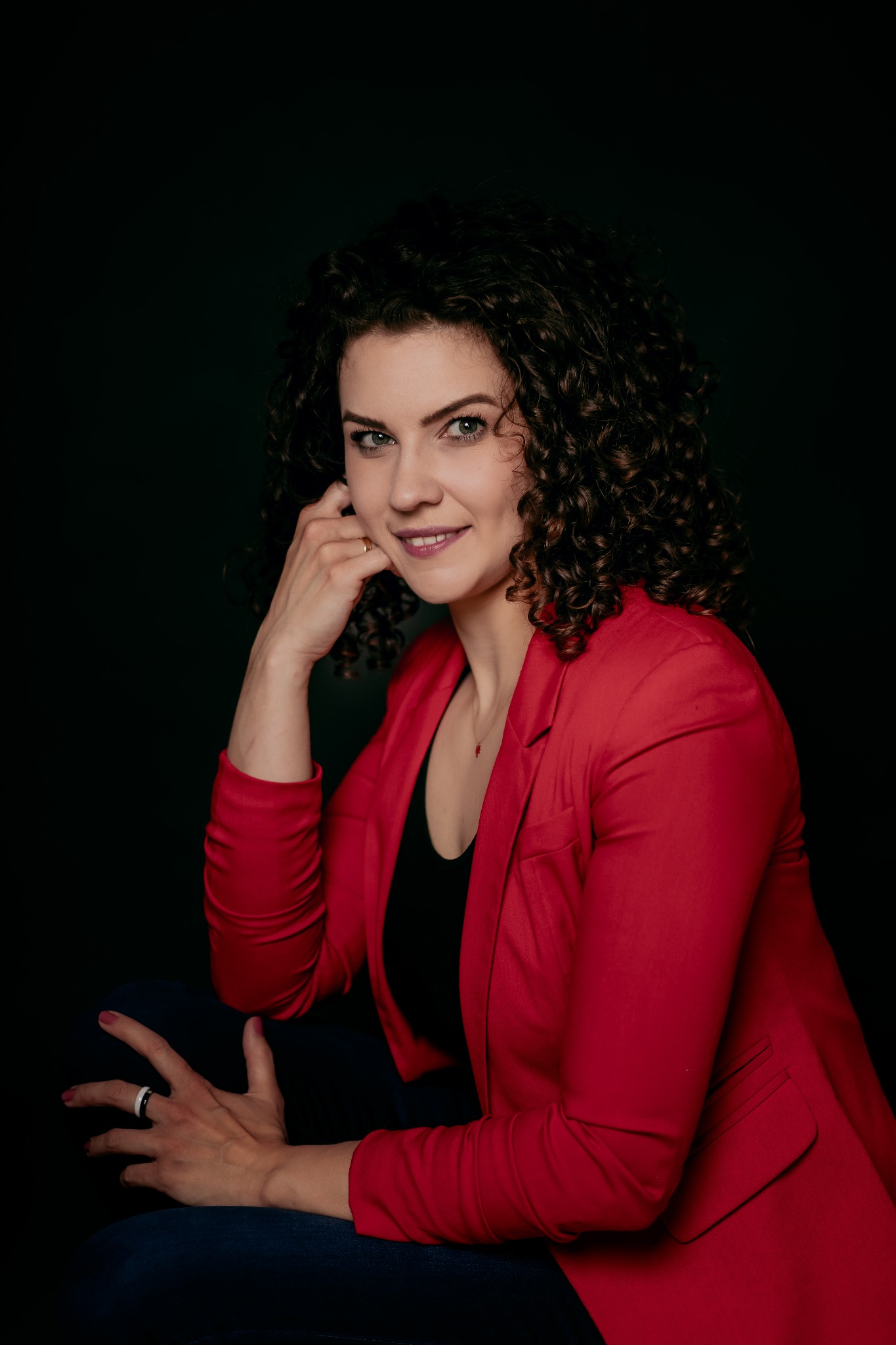 Uśmiechnięta kobieta z kręconymi włosami w czerwonej marynarce, opierająca brodę na dłoni, z pierścionkiem na palcu, na ciemnym tle.