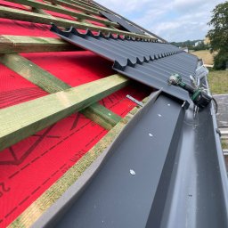 Dawid-Dach - Montaż grafitowej blachy trapezowej na dachu z widoczną membraną dachową w kolorze czerwonym i konstrukcją drewnianą, w tle zieleń i błękitne niebo, w kadrze wkrętarka akumulatorowa na dachu.