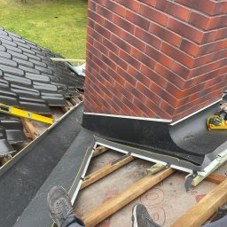 Dawid-Dach - Widok z góry na fragment dachu w trakcie montażu, z widocznym kominem z czerwonej cegły, poziomnicą, narzędziami i fragmentem nowej rynny, z butami osoby wykonującej prace w dolnej części kadru.