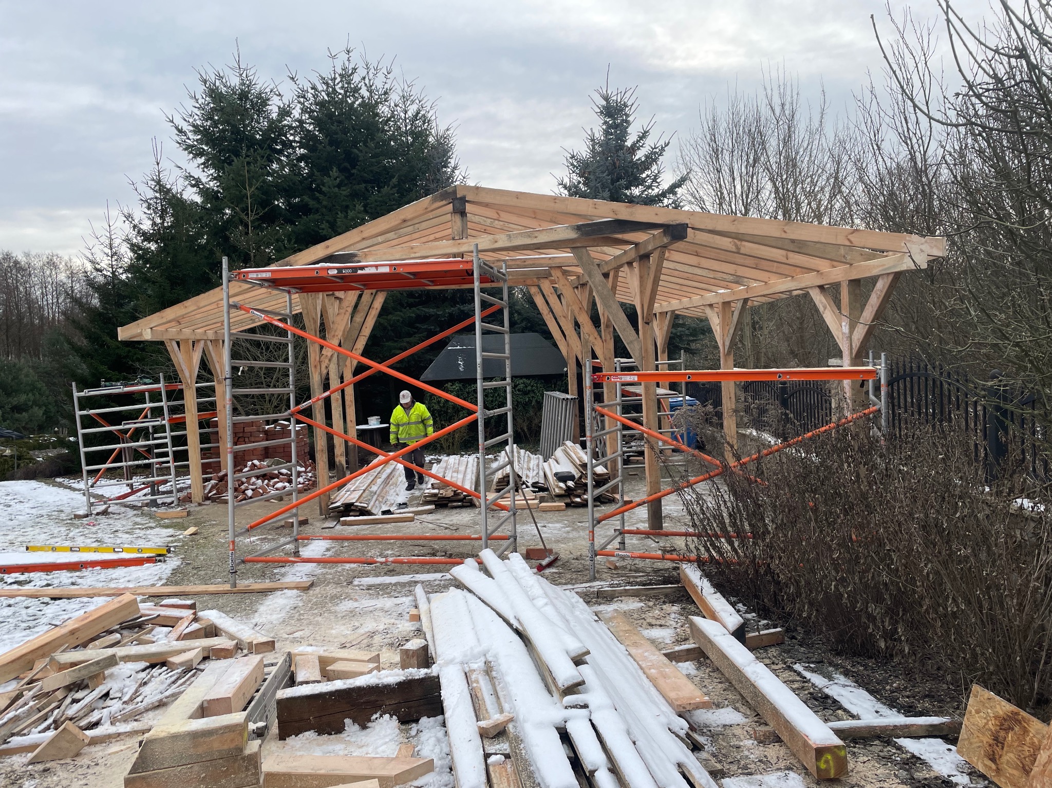 Konstrukcja drewniana zadaszenia w trakcie budowy, widoczne rusztowania, deski pokryte śniegiem, pracownik w odblaskowej kurtce w tle, zimowa sceneria.