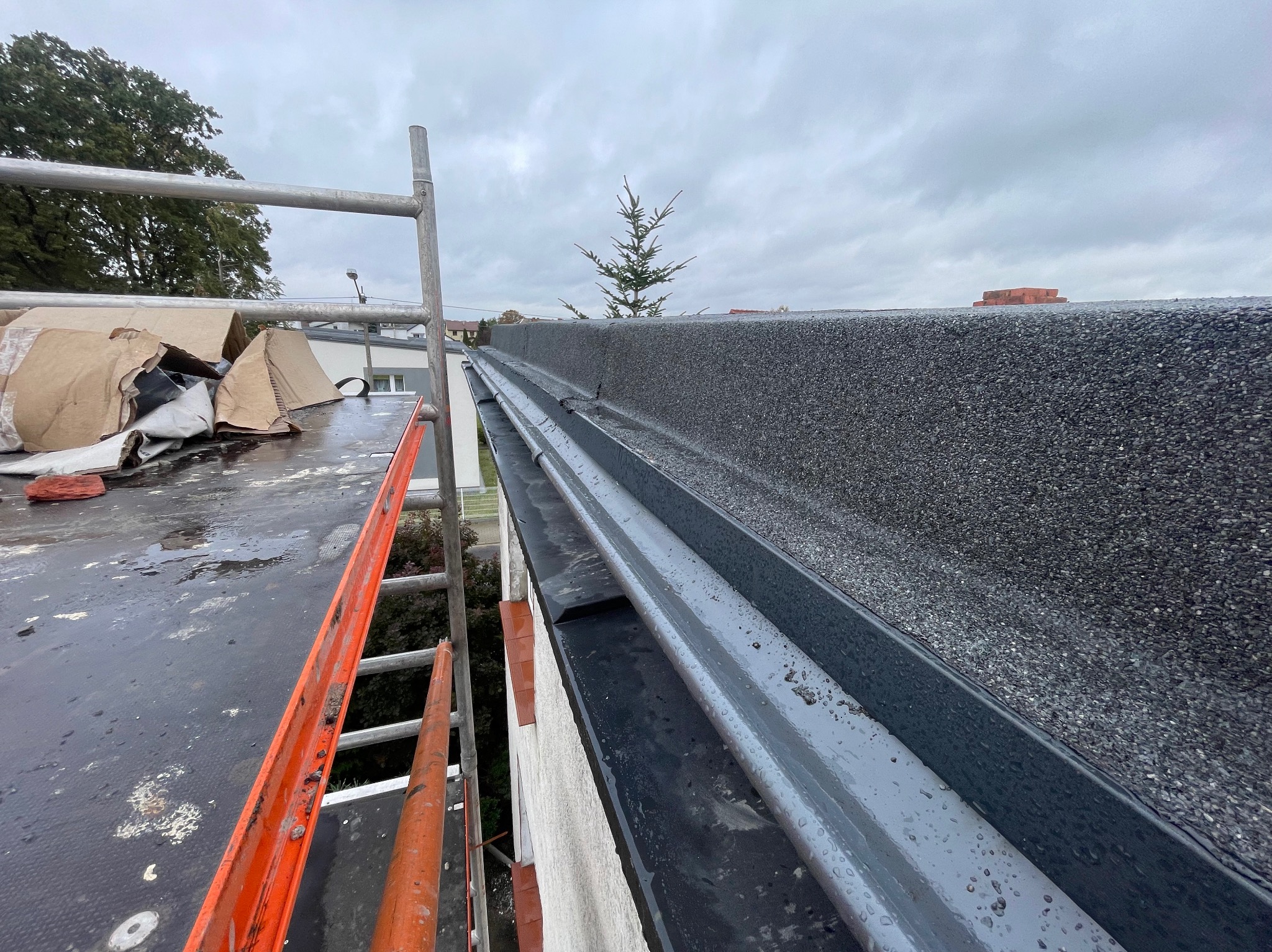 Widok z góry na płaski, bitumiczny dach w trakcie prac, z widoczną rynną, fragmentem drabiny oraz elementami uszczelniającymi na dachu, w tle pochmurne niebo i małe drzewko.