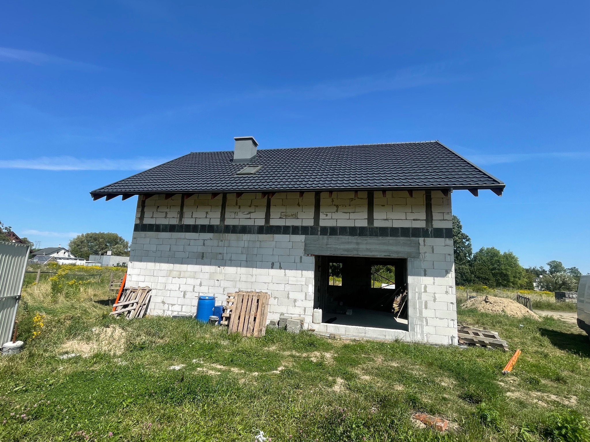 Surowy budynek w trakcie budowy z nowym, ciemnoszarym dachem z blachodachówki, widocznym kominem i niezadaszonym wejściem, na tle błękitnego nieba.