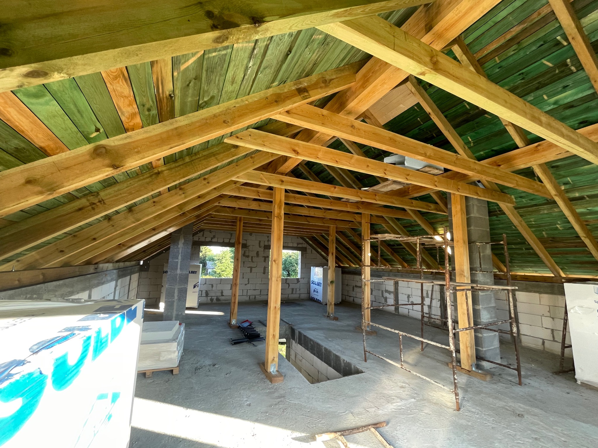 Wnętrze domu w trakcie budowy, widoczne drewniane elementy konstrukcji dachu z zielonymi deskami, ściany z bloczków betonowych i metalowe rusztowanie.