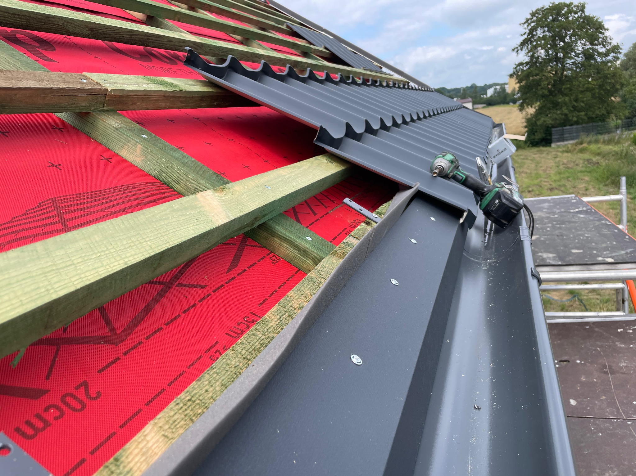 Montaż grafitowej blachy trapezowej na dachu z widoczną membraną dachową w kolorze czerwonym i konstrukcją drewnianą, w tle zieleń i błękitne niebo, w kadrze wkrętarka akumulatorowa na dachu.