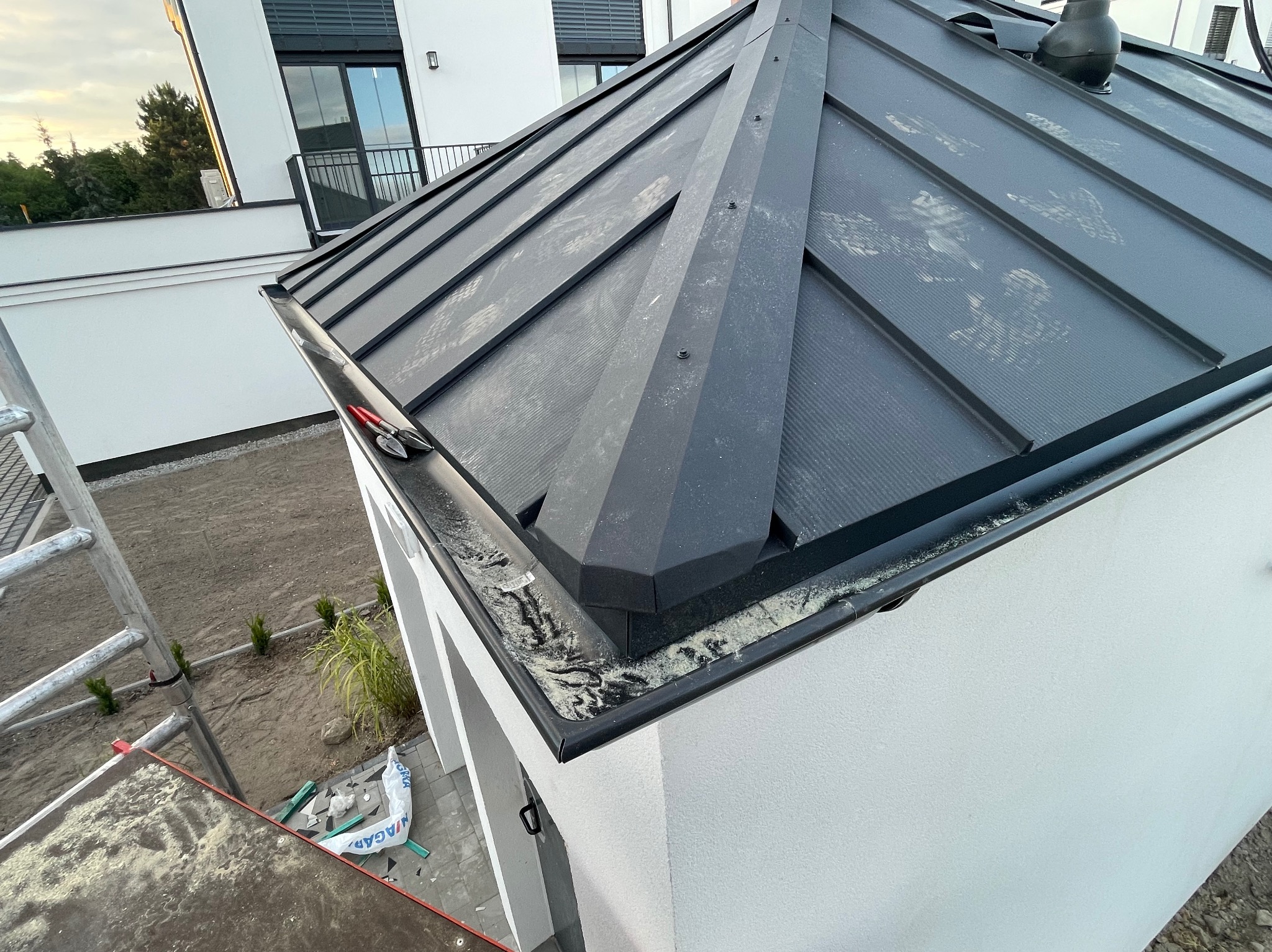 Nowoczesny, grafitowy dach z blachy na budynku jednorodzinnym, widok z góry na fragment dachu z rynną i narzędziami dekarskimi, ślady po pracach montażowych na powierzchni dachu.
