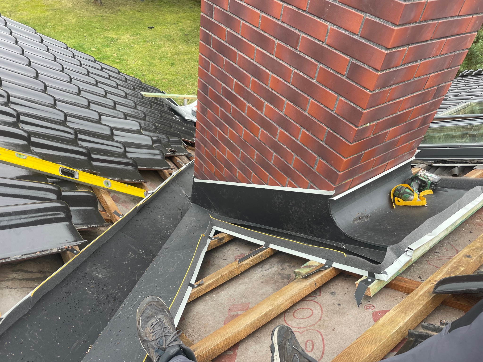Widok z góry na fragment dachu w trakcie montażu, z widocznym kominem z czerwonej cegły, poziomnicą, narzędziami i fragmentem nowej rynny, z butami osoby wykonującej prace w dolnej części kadru.