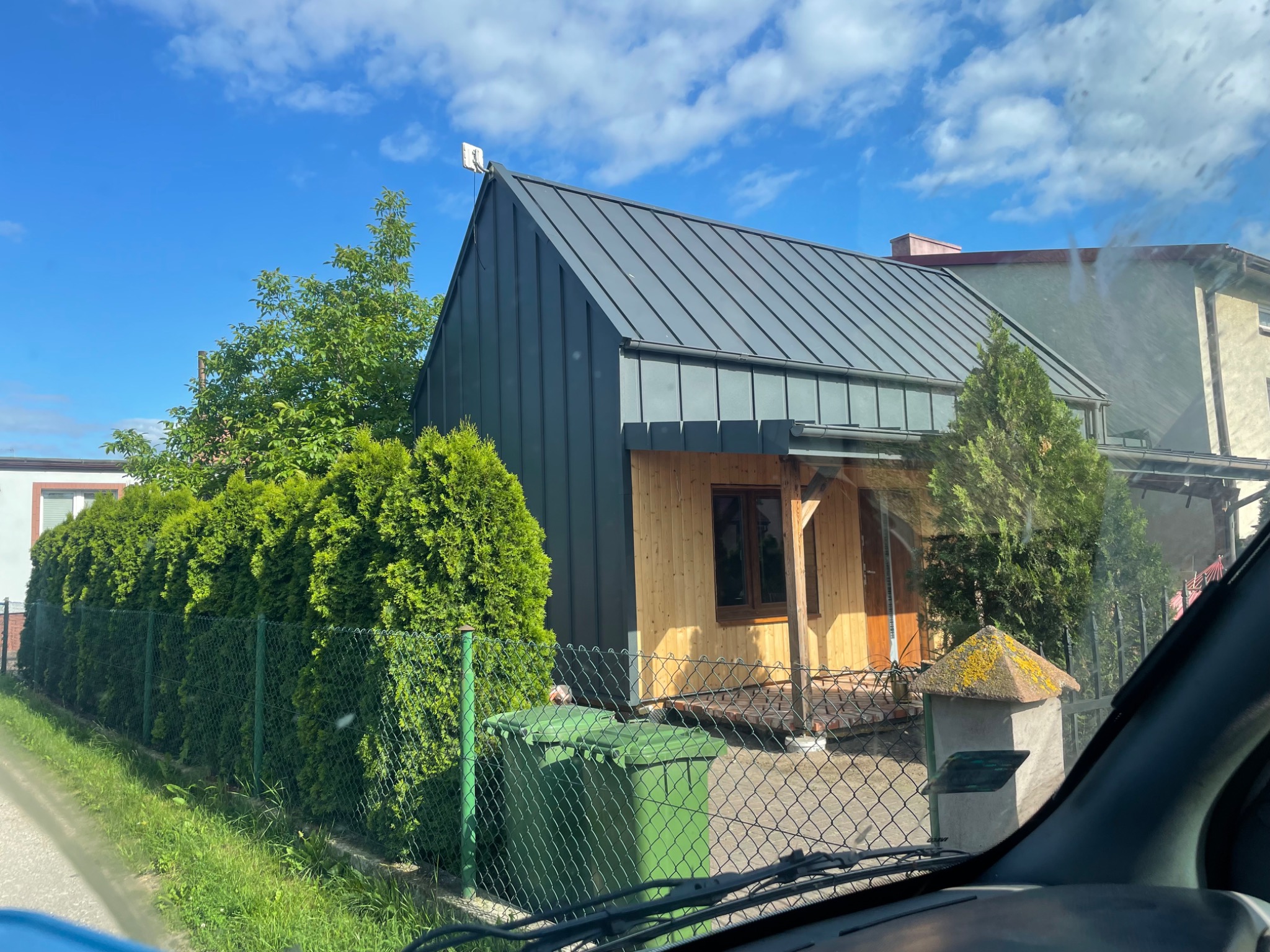 Nowoczesny dom szkieletowy z grafitowym dachem dwuspadowym, jasnym drewnem elewacyjnym i anteną na szczycie, widziany zza ogrodzenia i ulicy w słoneczny dzień.