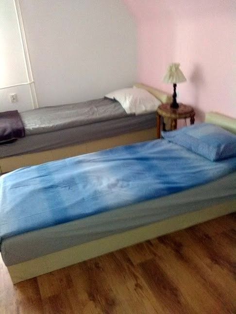 Pokój z dwoma łóżkami jednoosobowymi, jedno z niebieską pościelą, drugie z szarą, drewniana podłoga, lampka nocna na stoliku.