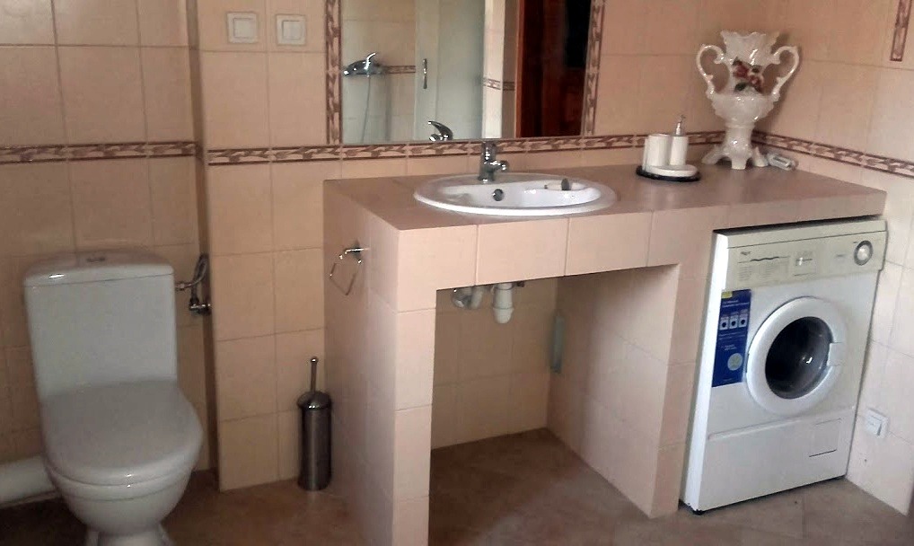 Łazienka z umywalką osadzoną na blacie z płytek, pod którym umieszczona jest pralka, obok toaleta, całość w beżowej kolorystyce.