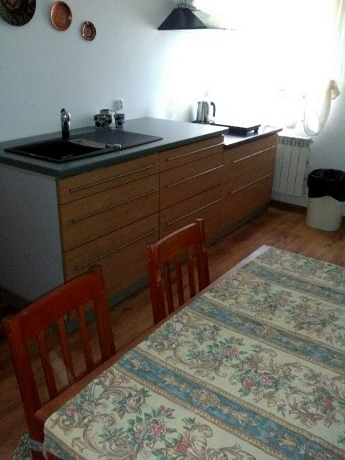 Wnętrze kuchni z widocznym blatem z szarym zlewem, drewnianymi szafkami z poziomymi uchwytami, stołem z kwiecistym obrusem i dwoma krzesłami.