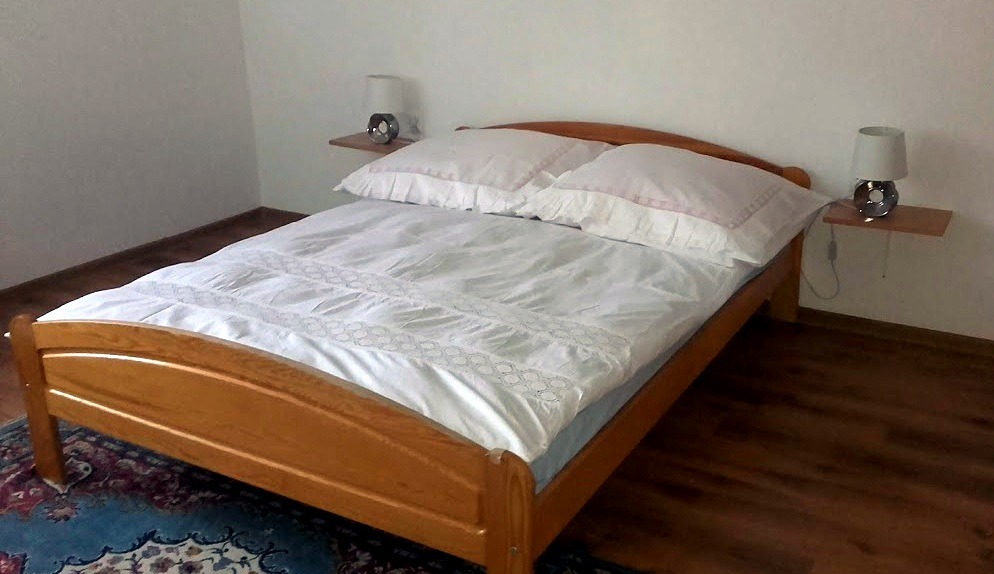 Jasne drewniane łóżko z białą pościelą i lampkami nocnymi na półkach w pokoju do wynajęcia.