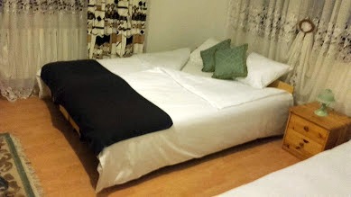 Pokój z łóżkiem dwuosobowym, nakrytym białą pościelą i czarnym pledem, z zielonymi poduszkami i drewnianą szafką nocną z zieloną lampką.