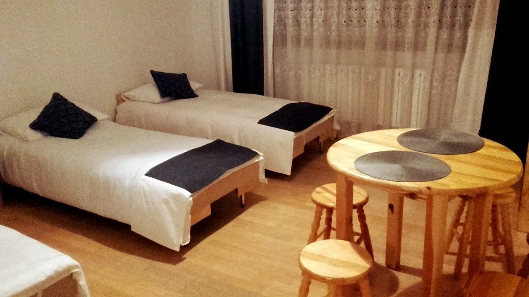 Pokój z dwoma pojedynczymi łóżkami z białą pościelą i ciemnymi narzutami, mały drewniany stolik z trzema stołkami, ciemne zasłony w tle.