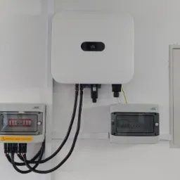 Biały falownik Huawei zamontowany na białej ścianie, poniżej dwie skrzynki elektryczne z czarnymi kablami, obok biała szafka z drzwiczkami i oświetlenie jarzeniowe.