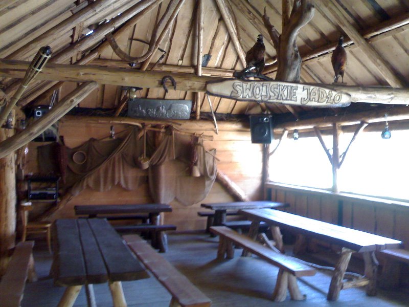 Rustykalna wiata z drewnianymi stołami i ławami, dekorowana trofeami myśliwskimi i napisem 'Swojskie Jadło'.