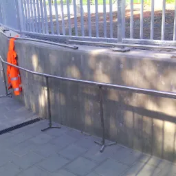 Metalowe barierki i poręcze zamontowane przy betonowej ścianie, na chodniku leży pomarańczowa kurtka robocza.