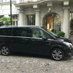 Czarny van Mercedes zaparkowany na brukowanej ulicy przed budynkiem z kolumnami i szklanymi drzwiami, prawdopodobnie hotel lub restauracja.