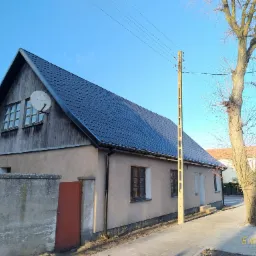 Dom jednorodzinny z ciemną, nową dachówką, anteną satelitarną na szczycie i drewnianą elewacją pod dachem, widoczny z chodnika w słoneczny dzień.