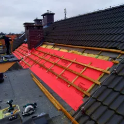 Rozpoczęty remont dachu z widoczną czerwoną membraną dachową Ventia Titanium Plus, drewnianymi łatami, częściowo ułożoną starą dachówką, narzędziami (wiertarko-wkrętarka, pilarka tarczowa Makita)...