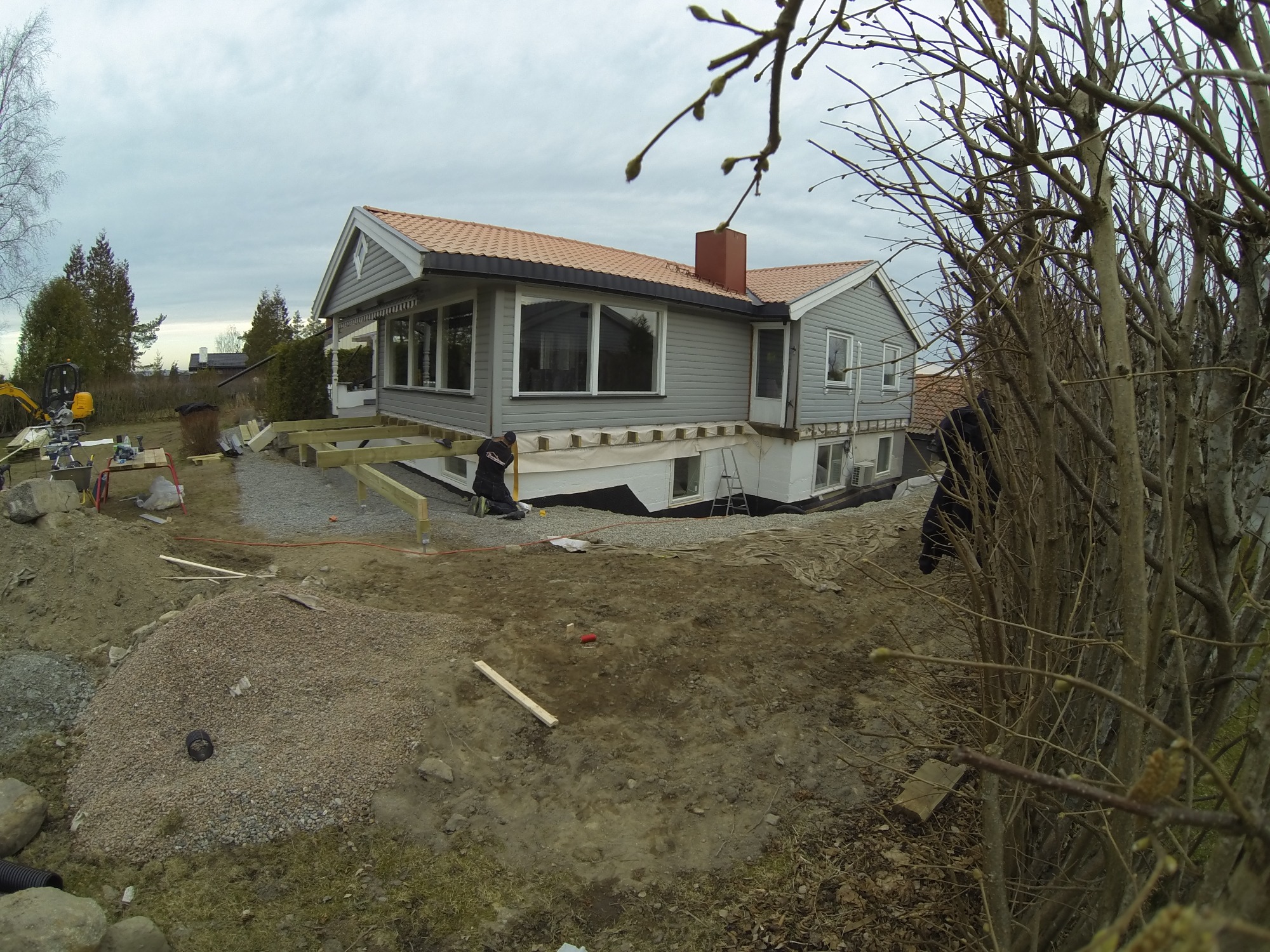 Szary dom jednorodzinny z pomarańczową dachówką w trakcie budowy tarasu, widoczny pracownik przy montażu drewnianej konstrukcji, w tle koparka i materiały budowlane.
