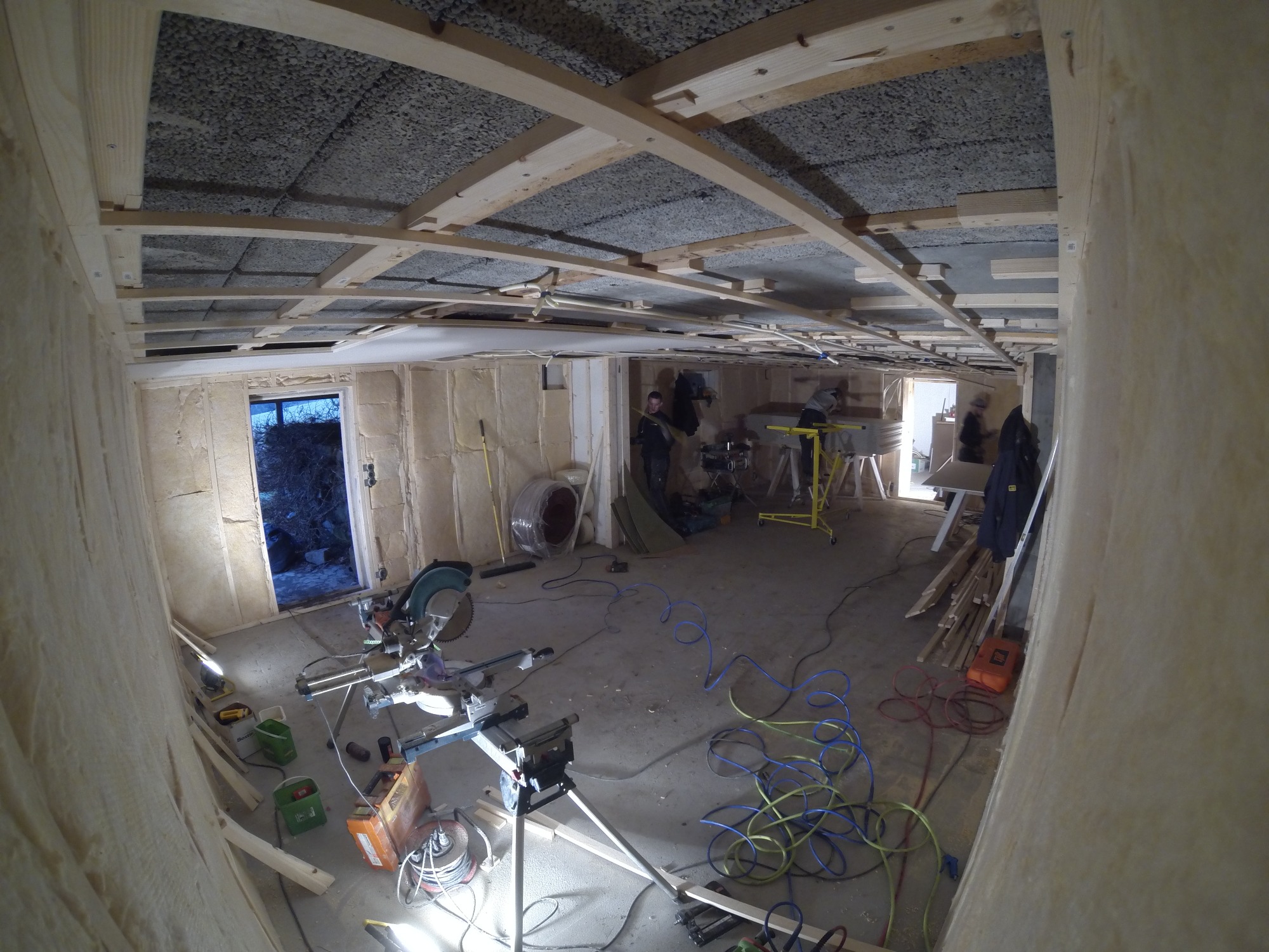 Wnętrze domu szkieletowego w trakcie budowy: widoczne ocieplone ściany, drewniana konstrukcja dachu z izolacją, narzędzia i ekipa budowlana w tle.