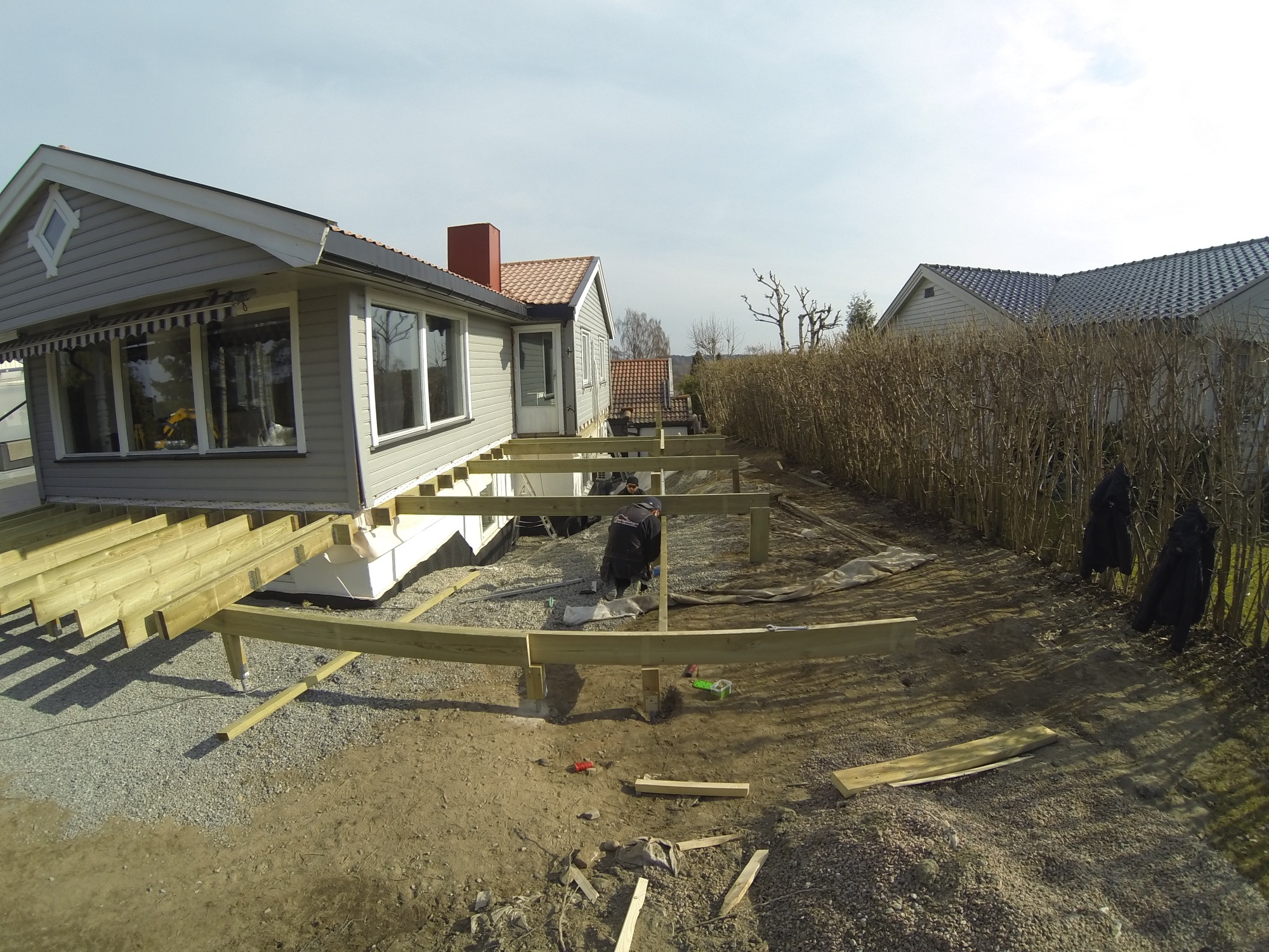 Montaż drewnianej konstrukcji tarasu przy domu jednorodzinnym, widoczne legary i słupy fundamentowe, w tle domy sąsiadów i krzewy.