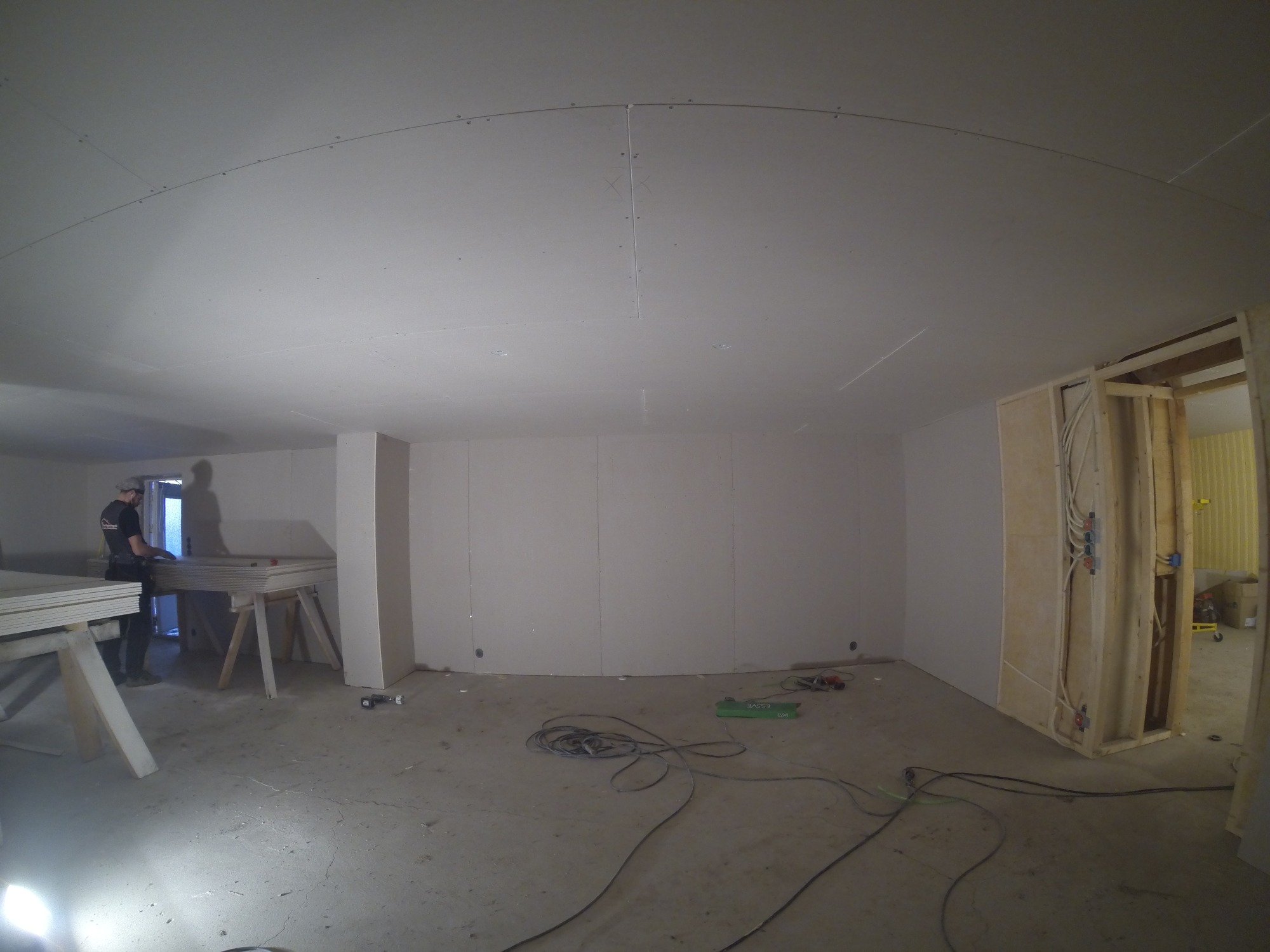Wnętrze w trakcie wykańczania, widoczne płyty gipsowo-kartonowe na ścianach i suficie z łukowym wykończeniem, elektryk pracuje w tle, przewody i narzędzia na podłodze.